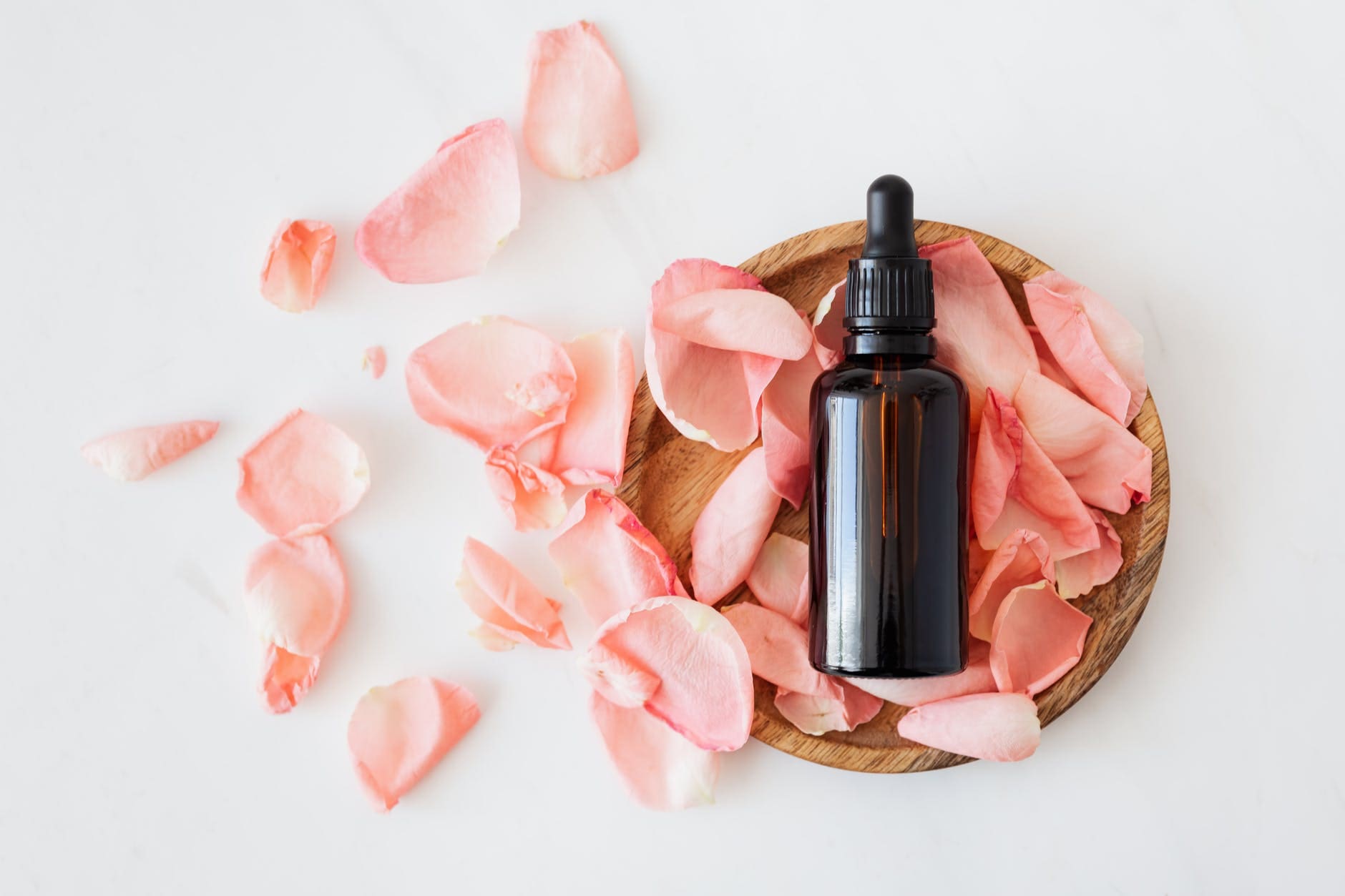 Glass bottles of essential oils arranged with pink rose petals and a wooden plate on a light background