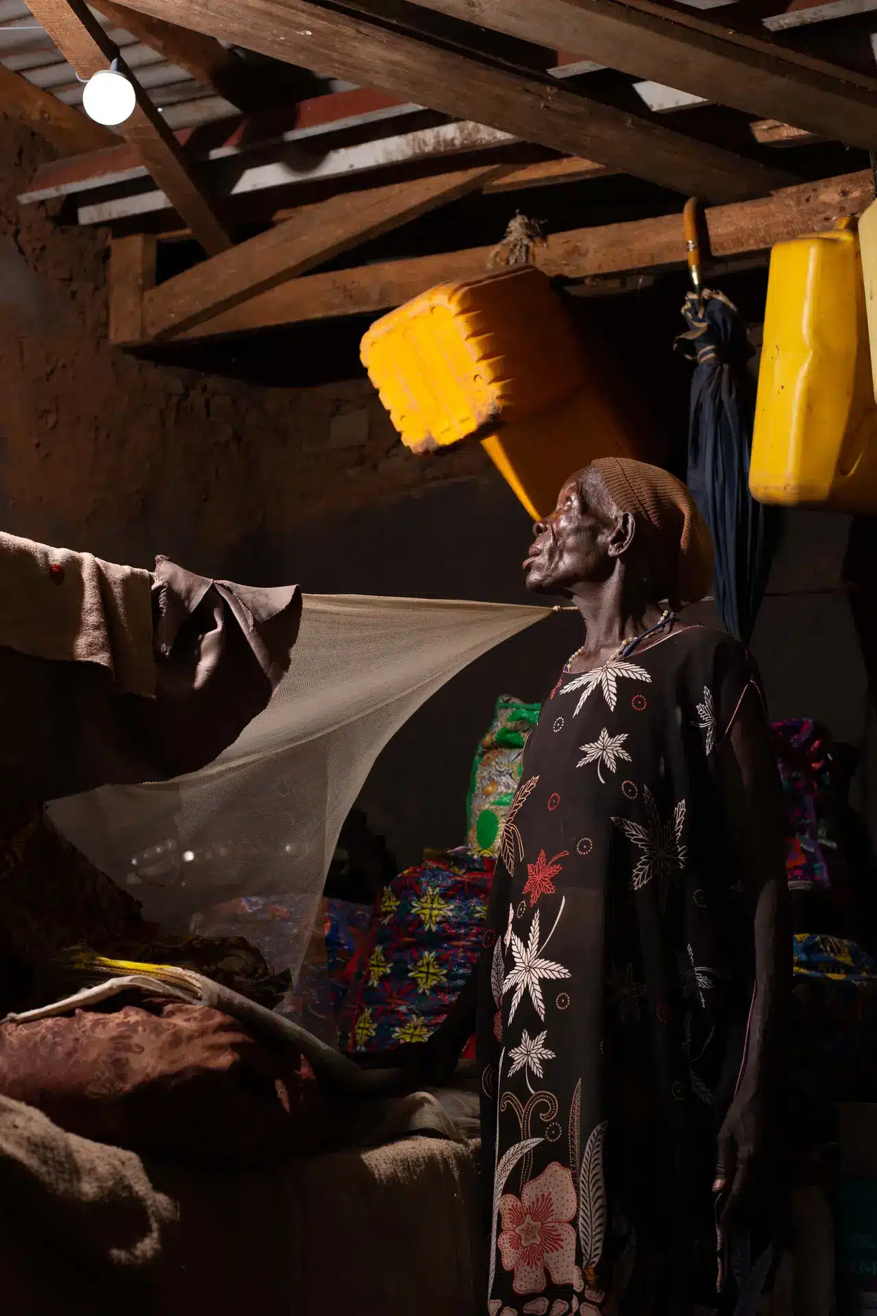 An elder woman in a dark floral kaftan spreads a mosquito net inside her home, lit from above by a solar-powered bulb, with yellow water jerry cans hanging from the rafters of a mud and timber structure in an Ubuntu Village partner community.