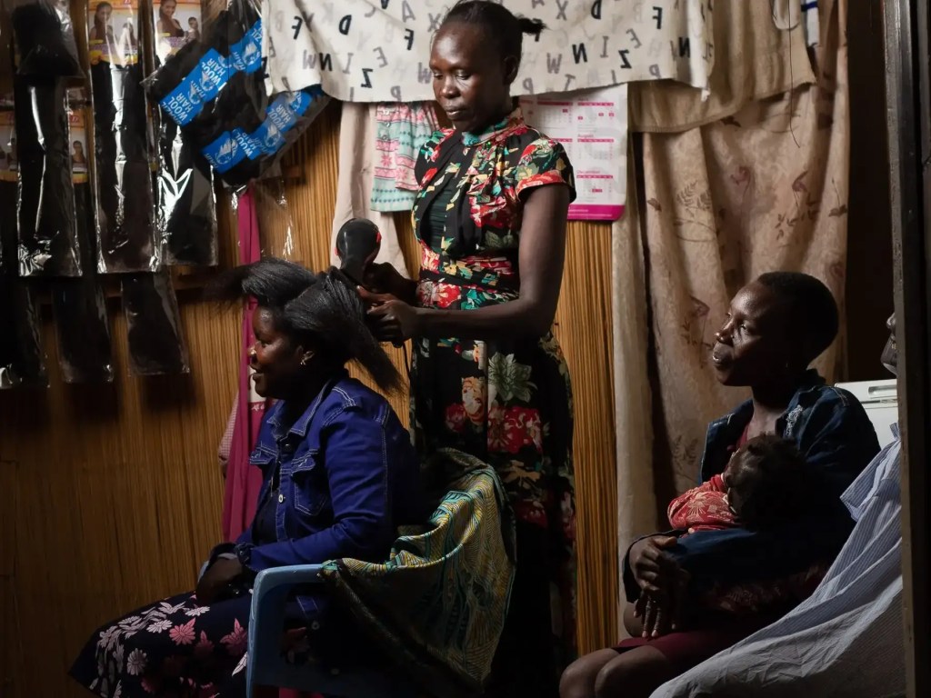 A woman in a floral dress styles another woman's hair inside a small salon lit by a solar-powered bulb, while a third woman sits nearby nursing a baby, all illuminated by clean energy in an Ubuntu Village partner community.